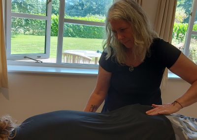 A woman performs spinal flow technique on a person lying face down in a room with large windows overlooking a green garden.