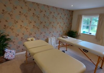 Two massage tables are set up in a room with floral wallpaper, carpeted floor, potted plants, and large windows letting in natural light.
