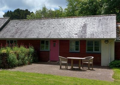 A small red cottage with a pink door, three windows, and a patio with a round table and two chairs, surrounded by greenery and trees.