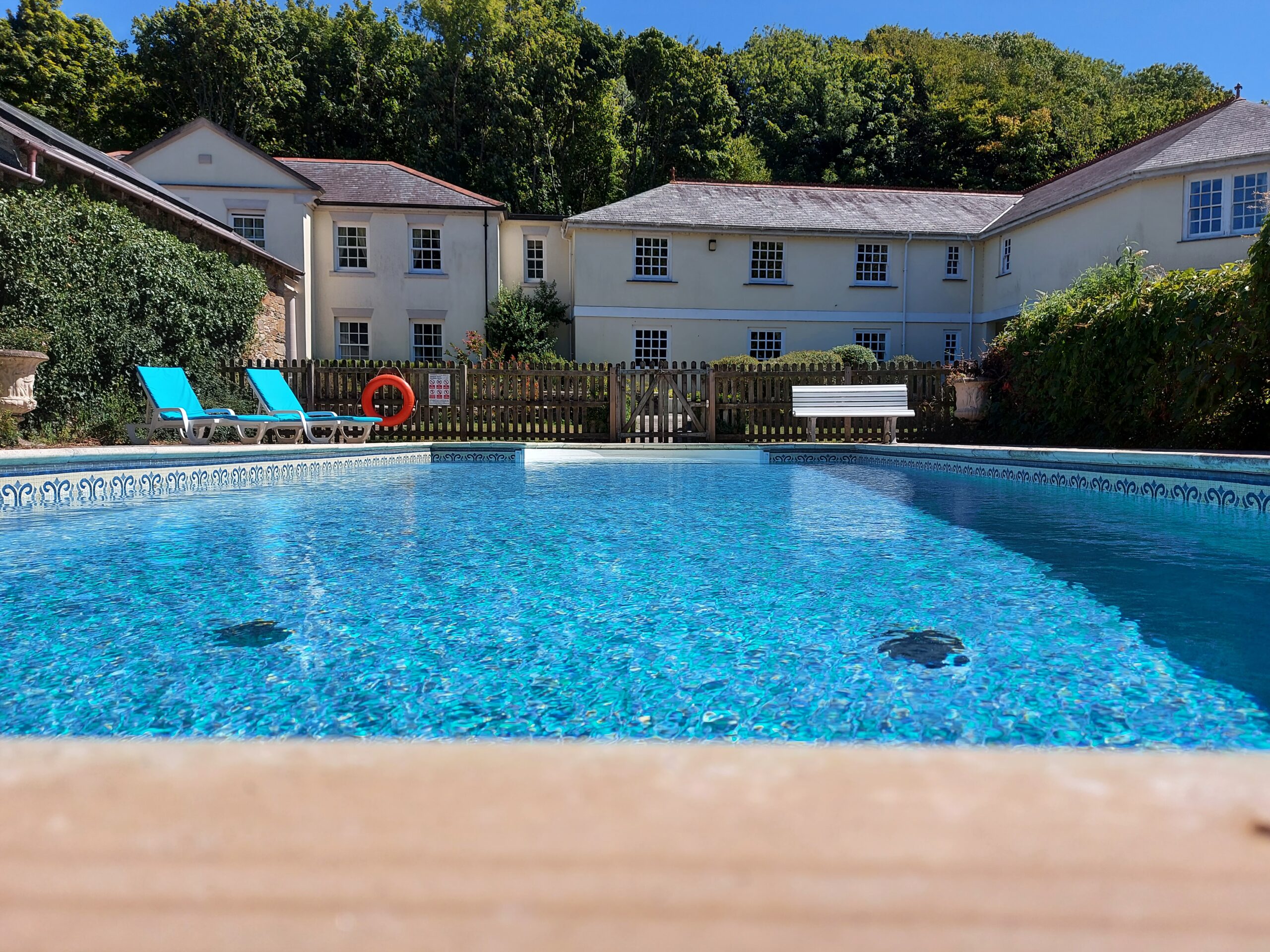 Outdoor swimming pool with clear water, surrounded by sun loungers and a building in the background on a sunny day.