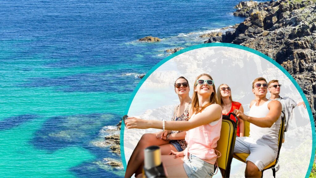 Cornwall-by-bus Five young adults sit together smiling and enjoying a scenic view of the rocky coastline and clear blue ocean under bright sunlight.
